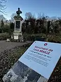 In the foreground is a sign in French that says Louis Braille monument and in the background is the nine foot tall monument, topped with a bronze bust of Louis.