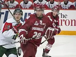 A group of three ice hockey players skate to the right of the image while looking to the left. The image is focused on the middle player, who is wearing the same uniform of the skater to his right, who is partially concealed by his teammate. Their opponent is to the left, with his team bench visible in the background.