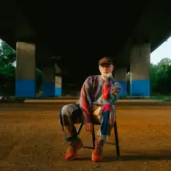 Ed Tullett wearing a colourful sweater and pants, with brightly coloured runners and a cap, sitting on a folding chair under a viaduct, head propped up on one hand, looking directly at camera
