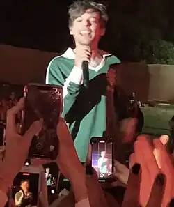 A man wearing a green sweatshirt singing into a microphone at a concert. Fans can be seen recording him with phones.
