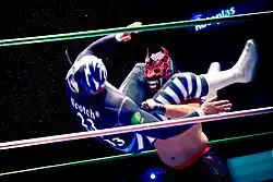 A masked wrestler executing a flying headscissorts, wrapping his legs around the head of his opponent, trying to throw him to the ground.