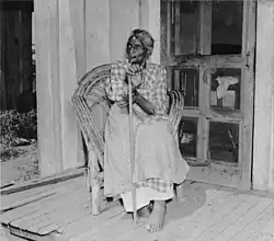 Black-and-white photograph of an elderly African-American woman sitting in a wicker chair on a porch. She has short hair and is wearing a checkered dress and an apron; she is barefoot. She is holding a cane in her hand and looking towards the left of the picture.