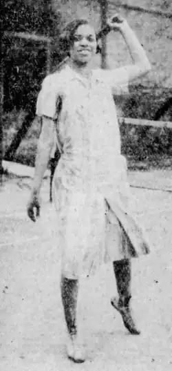 A young Black woman in tennis whites, on a tennis court, holding her racket overhead