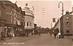 Photo Postcard showing a street scene of some shops, theatre, people walking, in cars and on bicycles and some tram tracks and overhead wires.