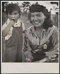 On the left is a small child aged approximately 4 or 5, wearing a white shirt and overalls. On the right is a smiling woman with shoulder-length dark hair. She is relaxed, crouched down next to her child. She is wearing a fringed-style buttoned shirt. The small child's hand is on her mother's left shoulder. They are standing outdoors , with a row of trees behind them in the distance.