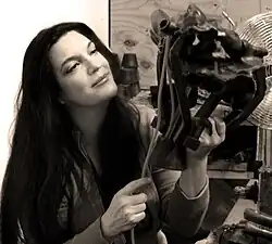 A woman with long dark hair looks thoughtfully at a sculpture she is working on