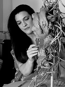 A woman working on a clay sculpture of a face attached to a metal framework, focusing intently as she refines the details with a sculpting tool