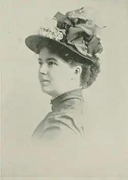 19th-century B&W portrait photo of a woman with her hair in an up-do, wearing a dark, fitted blouse and a hat.