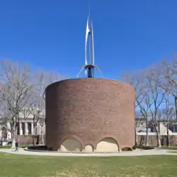 An aluminum spire tops a simple textured brick cylinder, surrounded by a reflective moat