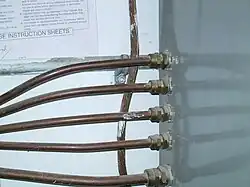 Five metal-covered cables enter the left side of an electrical panel through brass fittings, with bare cable running down the concrete wall behind.