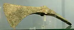 Verdigrised pickaxe. The bottom edges of the axe blade and the pick form one continuous curve. The top of the axe is also mildly concave between the eye and the blade. The sharp edge of the blade curves from nearly parellel to the (missing) handle to nearly at 45 degreees to it.