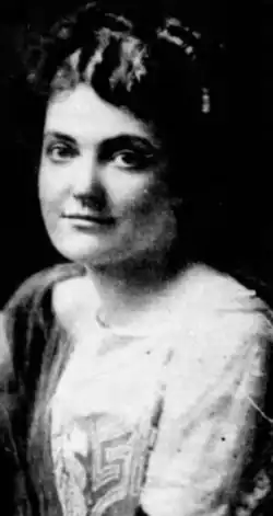 A young white woman with dark hair, wearing a light-colored dress with beaded embroidery on the bodice