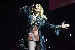 A blonde woman sings to a microphone. She's dressed in a brown corset underneath a leather jacket, and carries a guitar to her back.