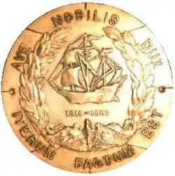 A round brass metal object features a sailing ship in the center over the U.S. Navy submarine service chest insignia of a dolphin and framed in a laurel wreath. In a circle around the edges are the Latin words "Ave Nobilis Dux, Iterum Factum Est" which translates to "Hail Noble Captain, It is Done Again."