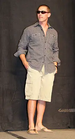 A man wearing cargo shorts, flip-flops, a buttoned shirt and sunglasses