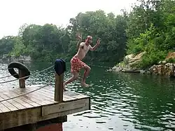 A man in a swimsuit jumps off a dock mid-air laughing into Beaver Dam.