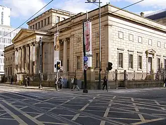 A neopalladian cuboid building as viewed centred on its corner from across the street. Halfway down the facade is a portico, and there is a central cuboid atrium that rises in the centre of the roof above the main roof height. There are regularly arranged sash windows in two stories.