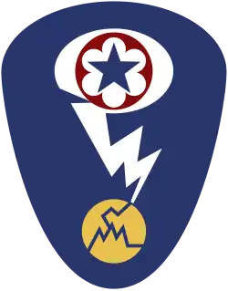 Oval shaped shoulder patch with a deep blue background. At the top is a red circle and blue star, the patch of the Army Service Forces. It is surrounded by a white oval, representing a mushroom cloud. Below it is a white lightning bolt cracking a yellow circle, representing an atom.