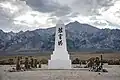 Photograph of a memorial at the Manzanar War Relocation Center. A low white obelisk with Japanese kenji characters and flower offerings stands against an expansive backdrop of the Owens Valley floor, snowy mountains, large clouds, and blue sky.