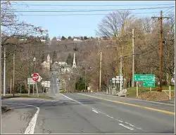A pair of highways meet at a signalized intersection. In the background at the base of a hill is a small village. One highway leads from the intersection, heads through the village, and ascends a larger hill in the distance.