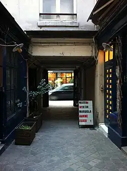 Narrow passageway with potted plants and lit windows leading to street with sandwich board sign reading "Maison Martin Margiela Homme Paris"
