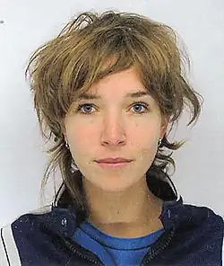 blurry image of Marike Jager wearing a blue t-shirt and dark blue and white jacket, looking directly at camera with slight smile