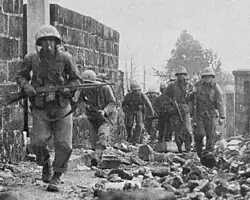 Group of U.S. Marines moving through rubble and debris in an urban combat area; the lead Marine moves forward while holding a Browning Automatic Rifle.