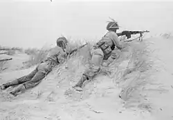 Black-and-white photograph of Dutch marines lying prone along a sandy ridge, aiming rifles outward during a military exercise.