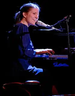 Markéta Irglová seen from profile wearing a dark blue dress and playing a piano on a darkened stage, looking sideways and grinning, face close to a microphone