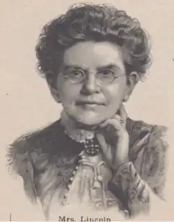 A white woman wearing glasses and a dark, high-colored dress or jacket; her dark, wavy hair is dressed in an updo