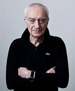 Elderly Vignelli pictured facing the camera, arms crossed, wearing a black, long-sleeved Izod polo shirt with an upturned collar.