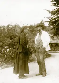 A woman dressed in black mourning clothes speaking outside with a bearded man wearing a hat.