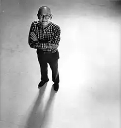 A bald man with glasses stands with arms crossed on a bright floor, casting a long shadow in front of him