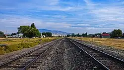 A pair of railroad tracks come from either side of the camera at the bottom corners to join into one midway us the image. On either side are two large grassy areas; on the right hand one a small wooden sign says "Melrose". There are streets with low buildings and some cars beyond the grassy areas, mountains in the distance, and blue sky with cirrus clouds above.