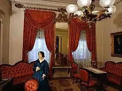 The front parlor, with various pieces of furniture. There is a mannequin in the foreground, a chandelier above, and two windows with curtains behind.
