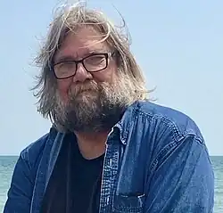 Schumacher in 2018, wearing his trademark denim shirt and black T-shirt, Lake Michigan.