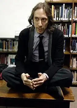 Michael Huemer sitting cross-legged on a desk, wearing dark pants and jacket, with a light-colored shirt underneath and a dark tie, fingers steepled in front of him, appearing to speak to someone to the right of camera, book-filled shelves in the background