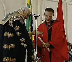Michael Elmore-Meegan standing at a lectern as an academic official confers an honorary degree during a formal ceremony