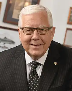 Photograph of a white man with white hair and glasses in a suit