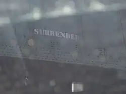 Miniature SURRENDER graffiti from 2007 stenciled on the green railroad bridge. Above the stencil are dark green squares where the previous graffiti has been painted over.