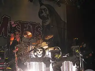 The rock band The Misfits performing onstage. The band's name in large lettering is printed on a fabric panel behind the performers along with a skull image. From left to right are the electric bassist, drummer, and electric guitarist.