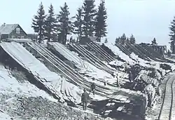 Men working next to chords of wood at the bottom of wooden ramps on the side of a hill. A building and set of trees is on top of the hill. A rail line parallels the chords of wood.