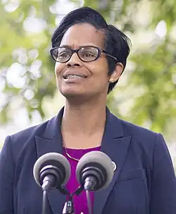 A black woman with short hair, and braces, wearing a navy blazer and purple shirt stands at a lectern.