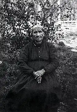Mother Solomon wearing a patterned dress and sitting in front of a bush with her hands in her lap