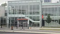 Photo of a glass station building with an entryway at ground level. An elevator and staircase are visible inside.