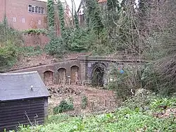 A buttressed brick retaining wall holds up the side of a railway cutting with the openings to a pair of tunnels on the right.