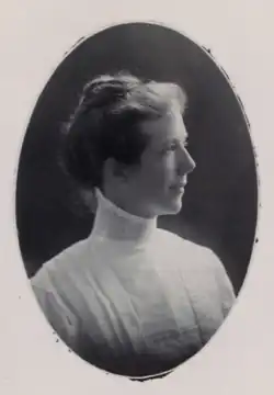 A white woman with dark hair dressed back, wearing a high-collared dark blouse or dress. She appears to have light eyes, and is slightly smiling.
