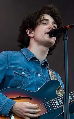 A poorly-shaved pale man with thick black curly hair with a microphone stand to his mouth wearing a jean jacket and holding a black and brown acoustic guitar standing in front of a black background.