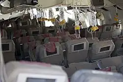 Interior of the aircraft, after the crash, showing oxygen masks hanging from the ceiling, ready for use. The seat backs have video displays installed, and most seats are in position, some reclined, some upright. The overhead compartments are open.