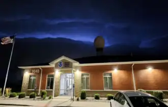 A National Weather Service office building, with an American flag shown on the left and address number "9040" shown on the office building. In the background, a water tower is shown in the middle of the office building. A lightning-illuminated shelf cloud is shown in the sky.